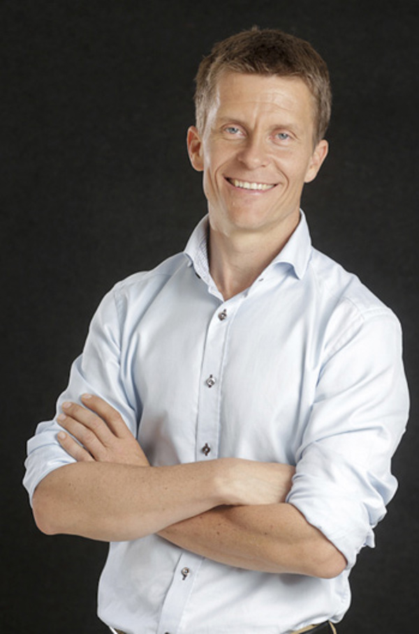 En smilende Ole Petter Hjelle står med armene i kors mot en svart bakgrunn. Han har kortklipt mellomblondt hår og lyseblå skjorte.