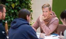 En gruppe studenter sitter rundt et bord. En mann i rosa hettegenser er i fokus. 