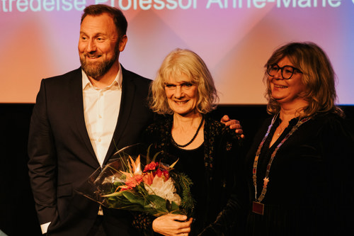 Anne-Marie Giørtz står i midten og smiler med blomster. På venstre side står em blid Kai Hanno Schmidt og på høyre side står prorektor Trine Johansen Meza og ser glad ut.