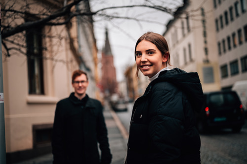 Two students standing in a street in Oslo, with a couch behind them