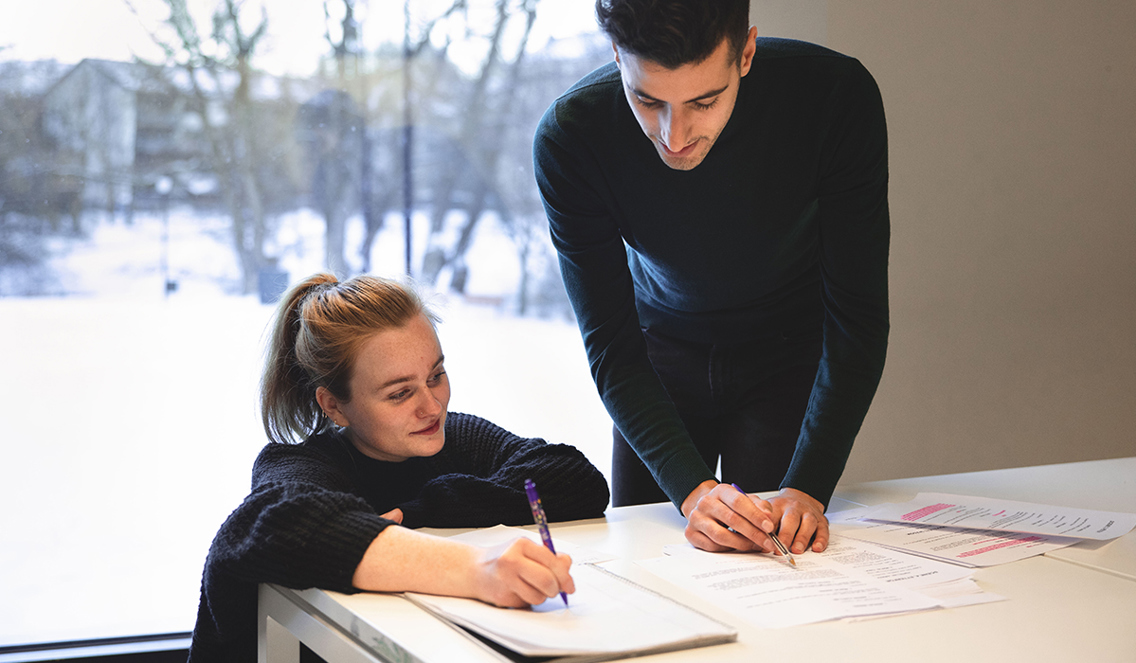 En jente står på huk, mens hun har den ene armen over bordet og skriver på et papir. Over henne står en gutt som ser ned på arket hun skriver på. 