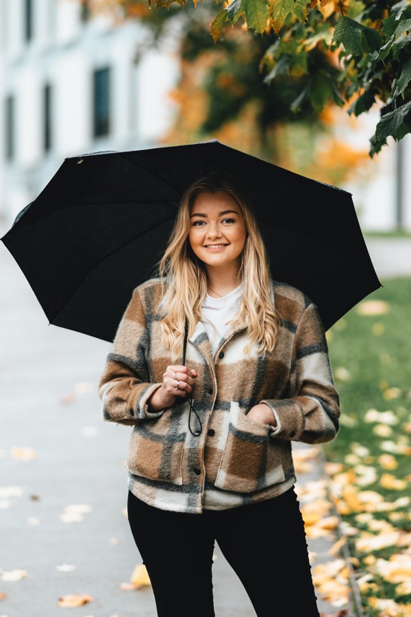 Bilde av ung blond kvinne med rutete høstjakke og paraply som står under et tre i regnvær.