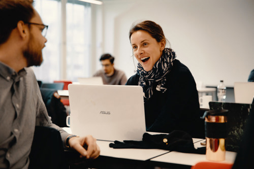 Studenter snakker sammen i klasserom. Den ene studenten har snudd seg rundt og ser på medstudent som sitter på pulten sin med en laptop.