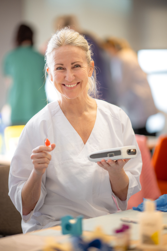 Person i hvit arbeidsuniform som holder et medisinsk instrument og en liten rød komponent under en praktisk øvelse