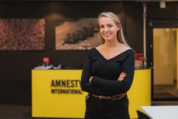 Foto av ung kvinne i sorte klær foran en gul stand med påskriften Amnesty.