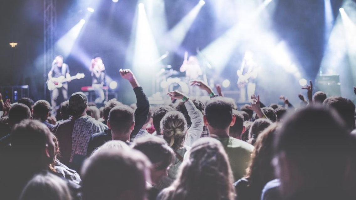  Bildet viser et publikum ved en konsert, vendt mot en scene med lysglimt og uskarpe musikere. En person i mengden løfter en arm, et tegn på feiring og glede. Atmosfæren er festlig og energisk.