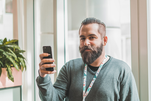 Foto av ung mann med skjegg foran et vindu som holder opp en telefon.