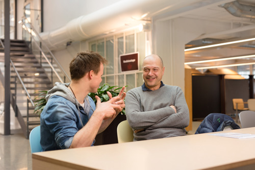 En mann sitter smilende ved at bord, og ser på en student som sitter med ryggen til oss.