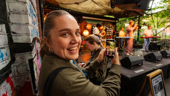 Student smiler inn i kamera mens hun filmer en konsert på mobilen