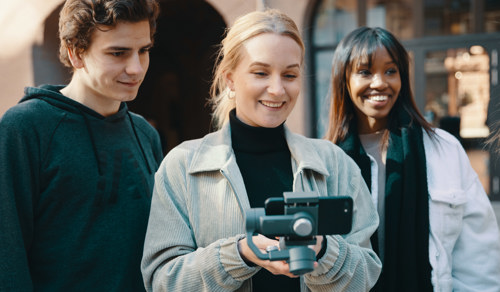 Student på Digital innholdsproduksjon, Maja Granli Jensen lærer seg å bruke mobil til å lage videoer. Hun har blondt hår og beige jakke, og filmer med mobiltelefon. Hun står med to klassekamerater, en gutt og en jente. De smiler og ser på mobiltelefonen mens de filmer. 