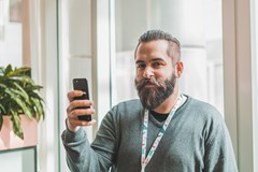 Foto av en mann med skjegg foran et vindu. Mannen holder opp en telefon.