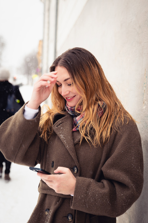 Portrett av Mina som står ute i snøvær, lent mot en vegg, mens hun ser ned på mobilen.