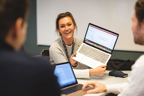 Foto av kvinnelig student som holder opp en laptop for å vise skjermen til to mannlige medstudenter.