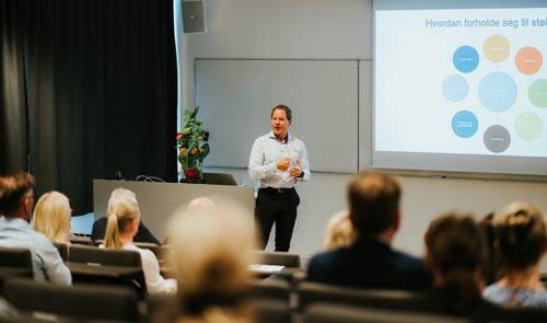 Kalhovde står foran i et auditorium og snakker til mange personer, som sitter med ryggen til. 