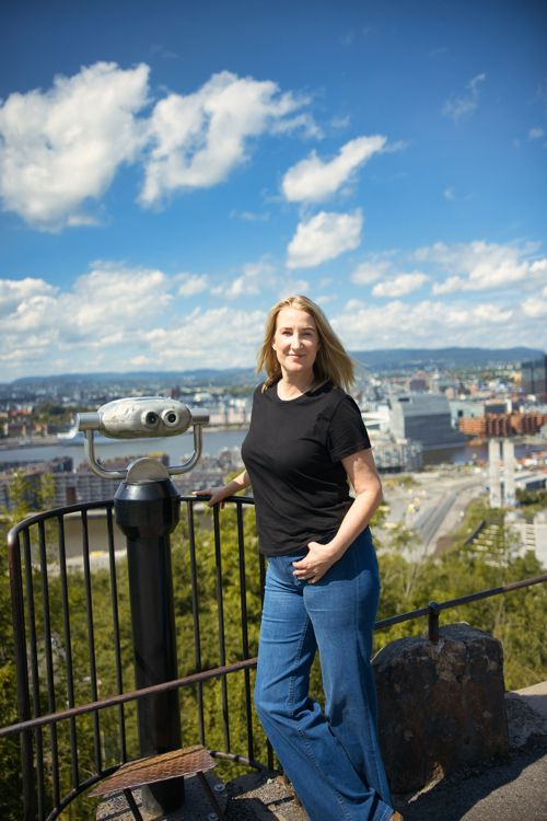 Christine poserer ved et utkikkspunkt ved siden av en kikkert, med utsikt over Oslo.