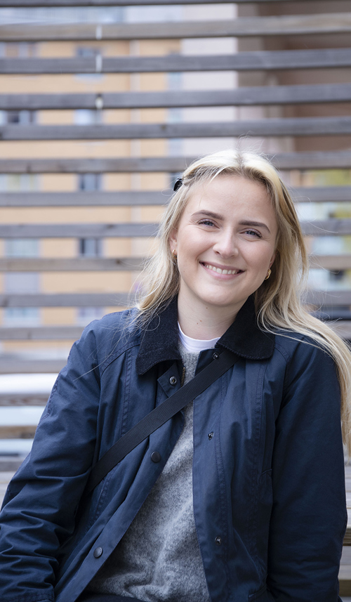 Kristiane Lunde studerer Music Business ved Fagskolen Kristiania. Hun sitter på ben benk med et brunt tregjerde i bakgrunnen utenfor inngangen til Campus Fjerdingen. Kristiane har langt, blondt hår og blå øyne. Kristiane har på seg en sort hårspenne, øredobber, en grå genser med en hvit topp under, en mørkeblå ytterjakke og har en sort stropp over skulderen. 