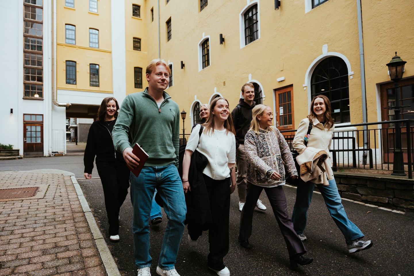 Studenter i gruppe går over plassen foran campusbygg i mur.