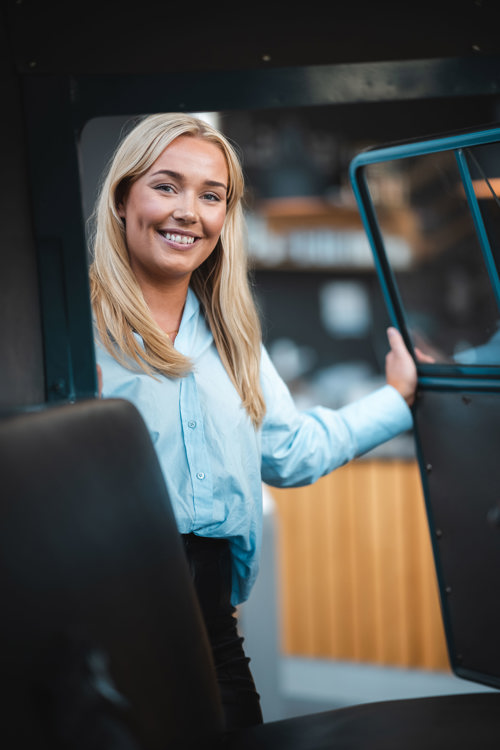 Blond dame i blå skjorte står ved åpen bildør.