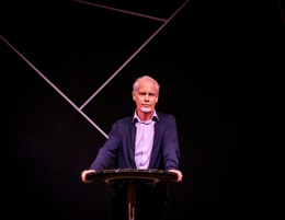Ingvard Wilhelmsen fotografert under et av hans populære foredrag. Han står på en scene og lener seg over et bord. Han har grått skjegg, blå cardigan og lyseblå skjorte.