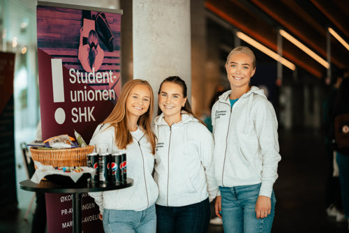 Tre jenter står og smiler til kamera på studenunionen stand