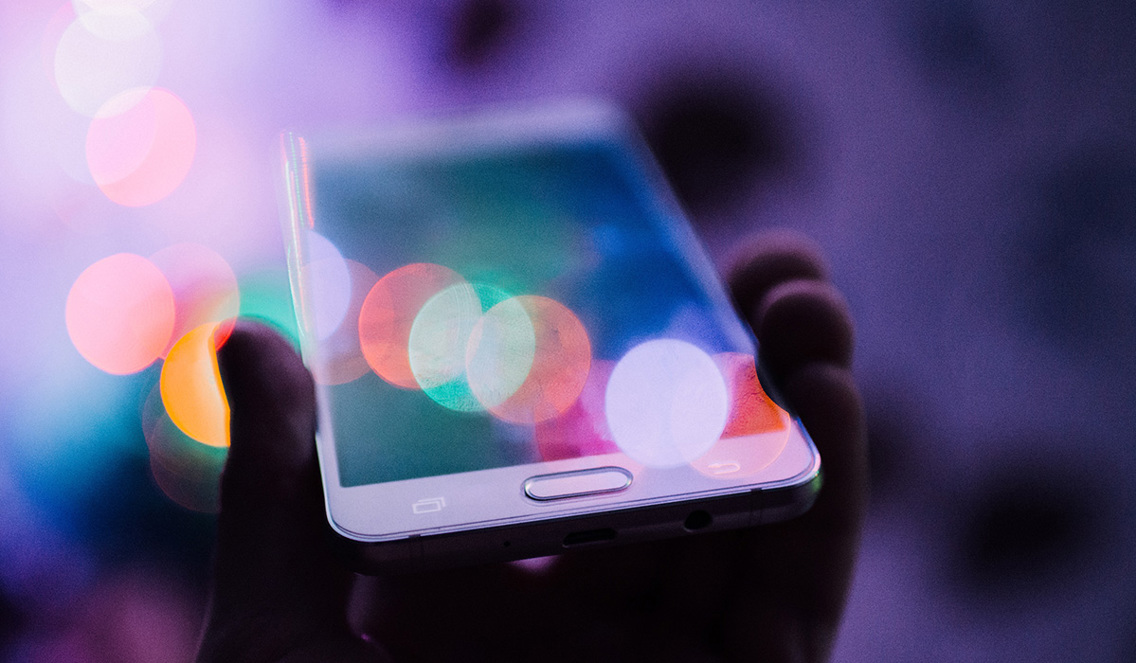 Bildet viser en hånd som holder en smarttelefon. Bakgrunnen er lilla og på skjermen kan man se små runde former i forskjellige farger som gjenspeilelse av lyset.