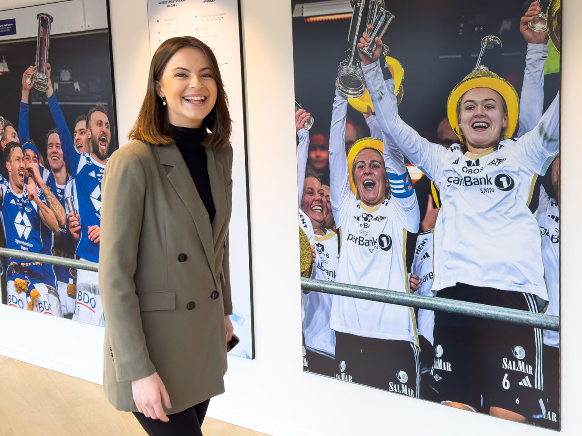 Smilende kvinne i blazer står i gangen på Ullevål stadion foran et bilde av spillere fra Rosenborgs kvinnelag som løfter cupgullet