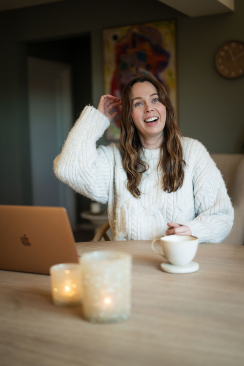 Monica sitter leende ved spisebordet med Mac og kaffekopp.