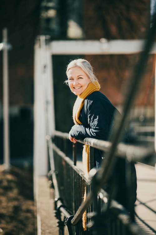 Voksen, blond dame i blå jakke og gult skjerf lener seg over et rekkverk på en bro.