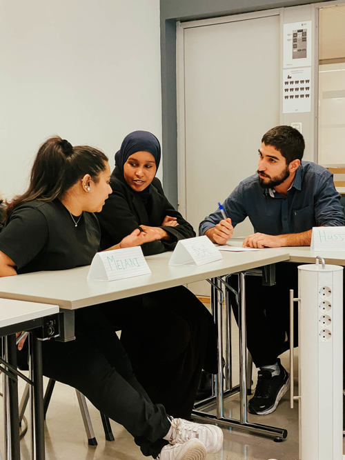 Tre studenter sitter ved en skrivepult mens de diskuterer seg i mellom. Det er en mann og to kvinner som samhandler. Foto.