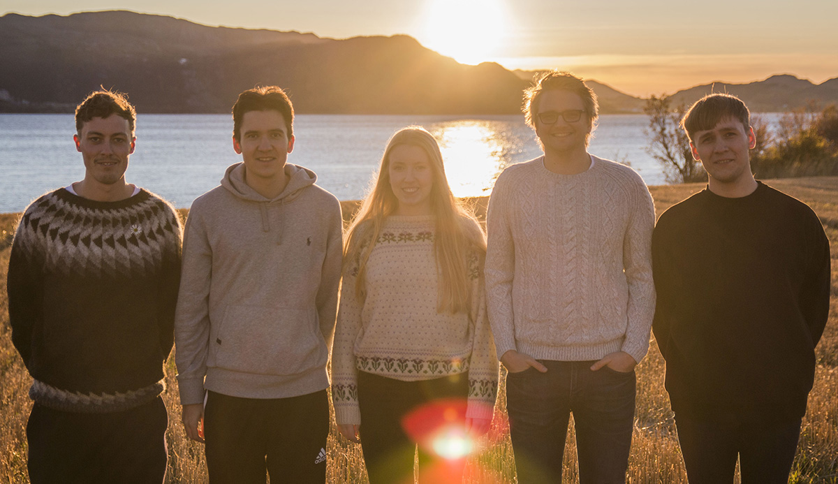 På rekke står de fem arrangørene å smiler til kameraet i en solnedgang med jorde, hav og fjell i bakgrunnen. FOTO