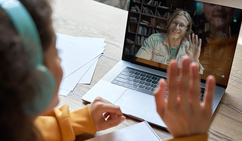 En kvinnelig student sitter ved et bord med en Mac fremfor seg, og har en videosamtale med en mann. De vinker til hverandre gjennom skjermen.  Kvinnen har blå hodetelefoner, en oransje genser og mørkt hår. Mannen på skjermen har blondt halvlangt hår, skjegg og briller.