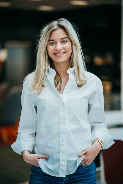 Ung, blond dame i hvit topp og blå jeans smiler til kamera.