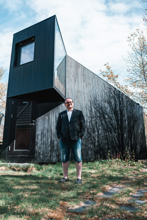 Mann i shorts og sandaler står utenfor en høyreist, arkitekttegnet skrivestue i hagen.