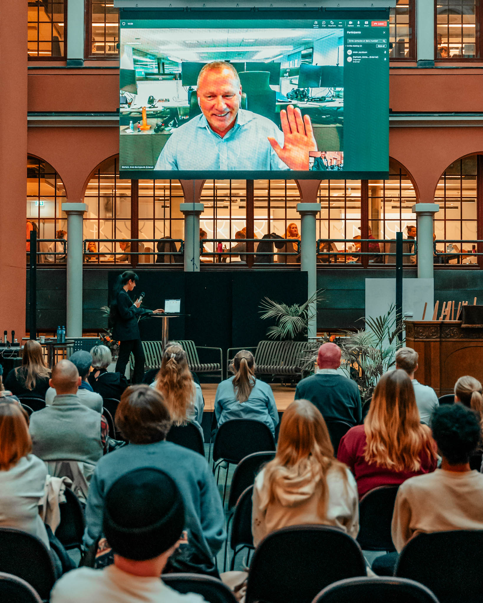 Nicolai Tangen sitter med en blå skjorte og hånden strekt opp til en hilsen. Han deltar live på en stor skjerm som ruver over forsamlingen. Han smiler blidt. Foto.