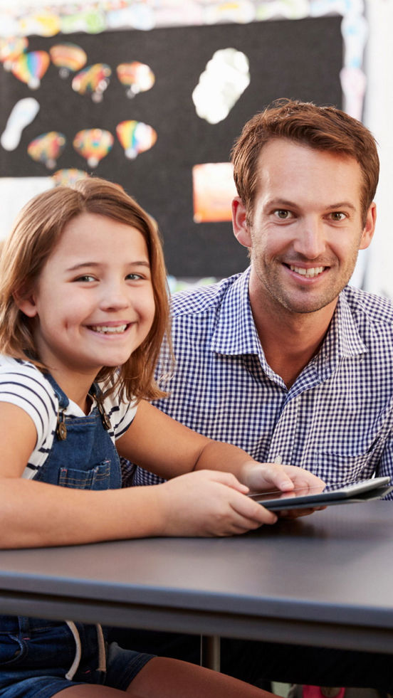 En smilende voksen og et barn ser på en nettbrett sammen i et klasserom. Bak dem er en tavle med fargerike kunstverk.