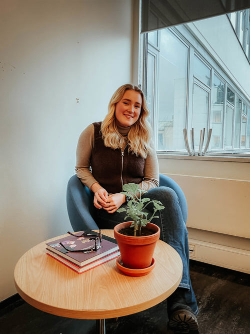 Selma Vedum Håland sitter i en turkis stol mens hun smiler mot kamera. Hun har hendene foldet til den ene siden og på bordet foran henne står det en liten potteplante. Hun har på seg strikkevest og jeans. Foto.