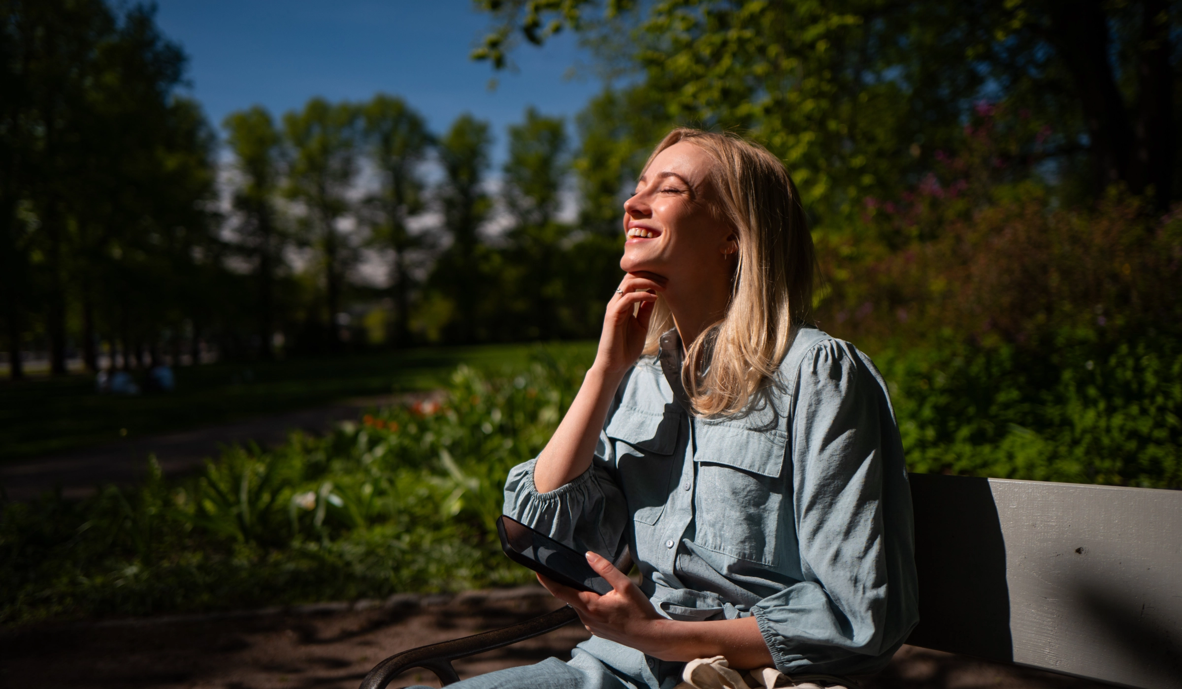 Blond kvinne sitter på parkbenk. Hun har telefonen i hånda, og ler ut i luften.