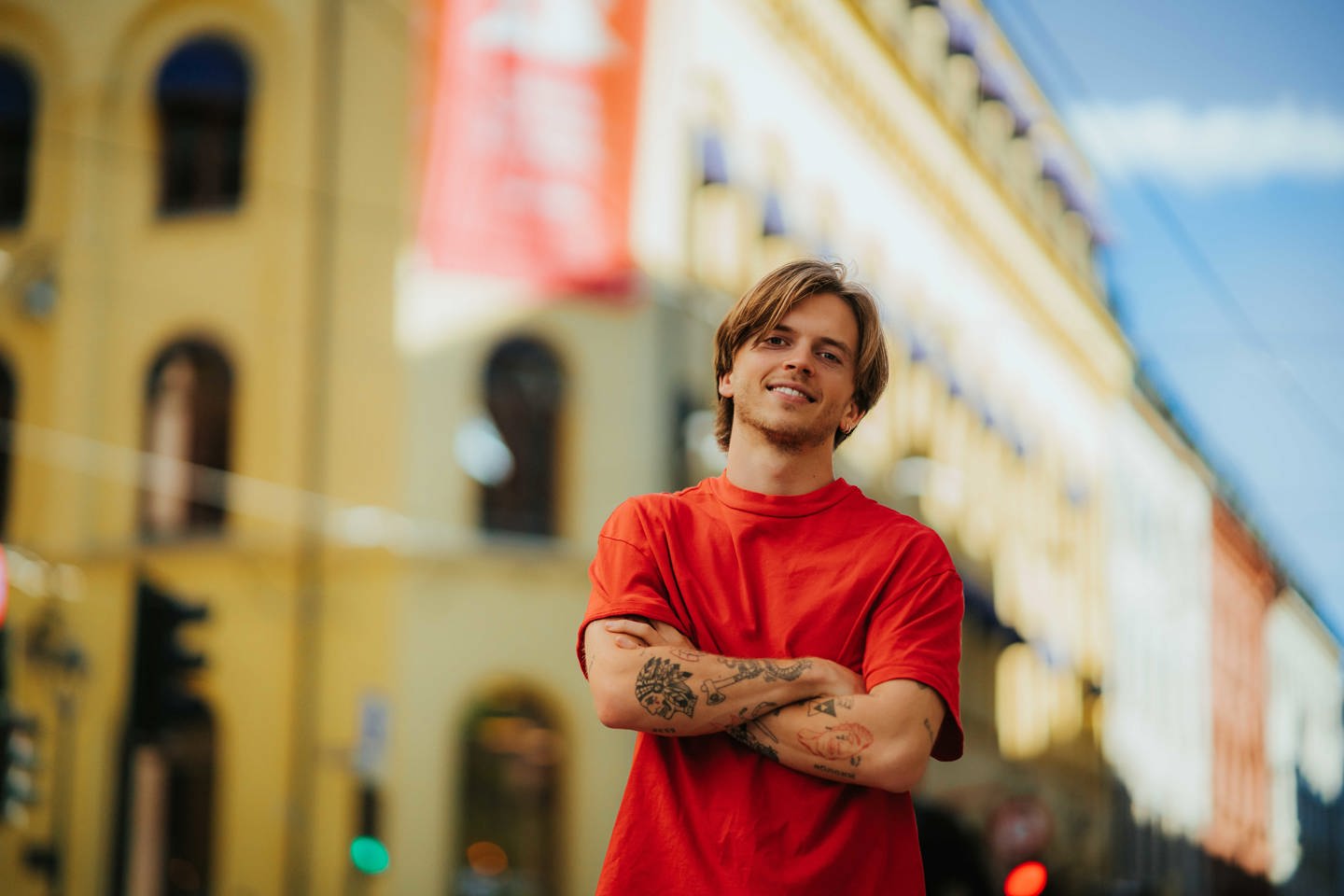 Bilde av en student med en lidenskap for kunst og design, står foran campus. Han har halvlangt brunt hår, tatoveringer og en rød t-skjorte.