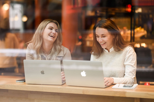 Cathrine og Helene sitter ved et vindusbord på en kafé med hver sin PC.