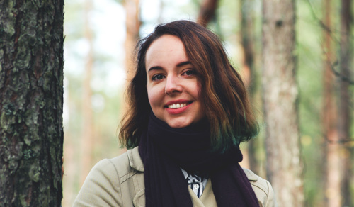 Cathrine Sandmæl er illustratør og har studert på Fagskolen Kristiania. Hun er i skogen og smiler mot kamera iført svart sjerf og beige jakke. Hun har brunt halvlangt hår. 