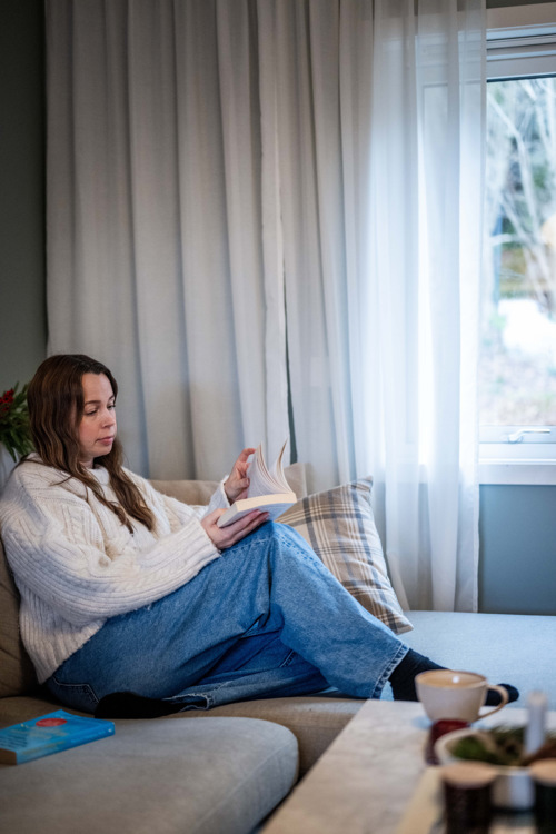 Monica sitter i sofaen hjemme og leser en pensumbok.