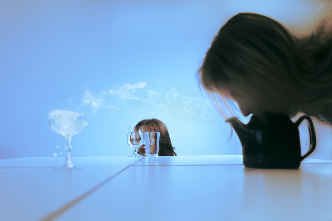 Foto av et glass med bobler, som avgir røyk, som igjen blåser mot en person på siden som blåser ned i en tekanne. I bakgrunnen ser vi en person som kikker gjennom to ulike drikkeglass. Lyset bryter gjennom glassene og bøyer og forvrenger ansiktet hens.