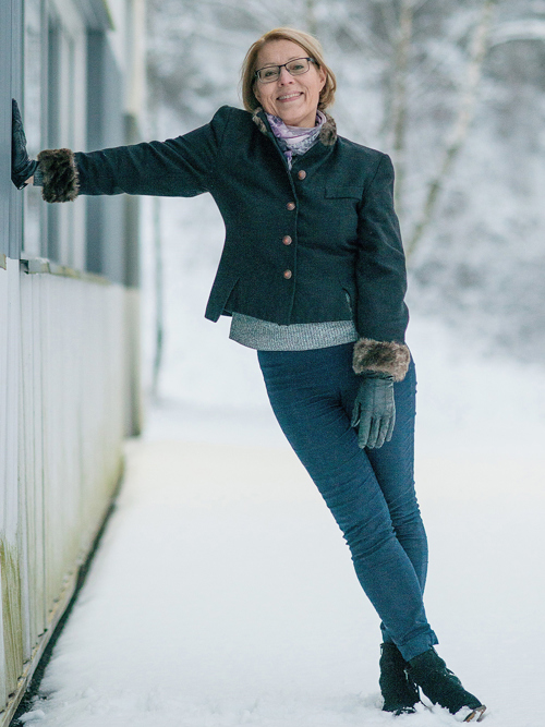 Eva Widenoja lener seg mot et bygg og smiler. Hun har blondt hår og har på seg blå jeans, grønn vinterjakke, skjerf, grå genser, svarte sko og skinnhansker. 