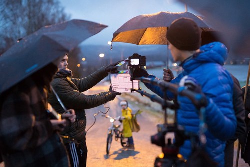 Studenter står ute og filmer i regnet med paraplyer. Foran en person på sykkel står en student med en klapper.