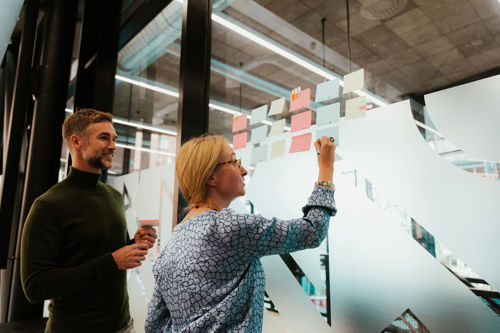 En mann og en kvinne til venstre i bildet. De vurderer en rekke post-it-lapper som henger på en glassvegg.