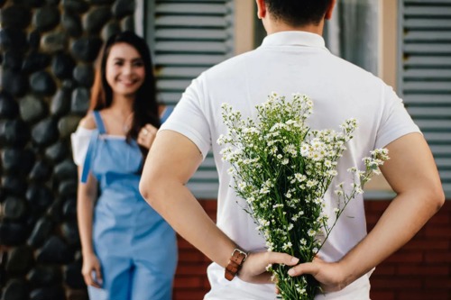 Kvinne ser mot kamera mens en mann står foran henne med ryggen til kamera og blomster bak ryggen sin.