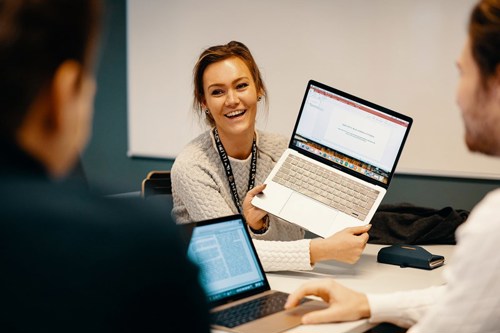 Foto av kvinnelig student som holder opp en laptop for å vise til to mannlige medstudenter.