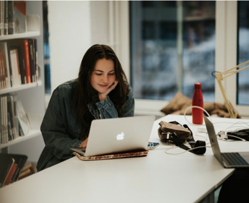 Kvinne som sitter foran Mac-en ved en pult i et bibliotek.