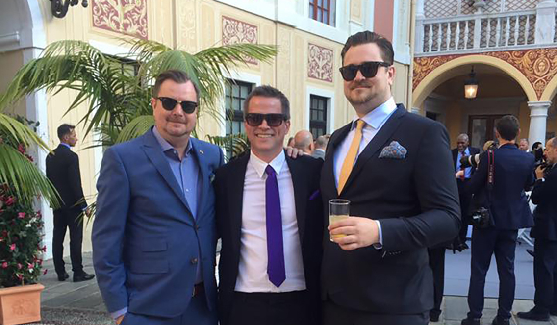 Tre menn I dress og solbriller, smiler og holder rundt hverandre. 