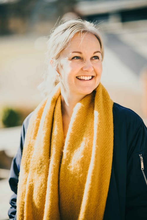 Voksen, blond dame med AirPods og gult skjerf smiler.